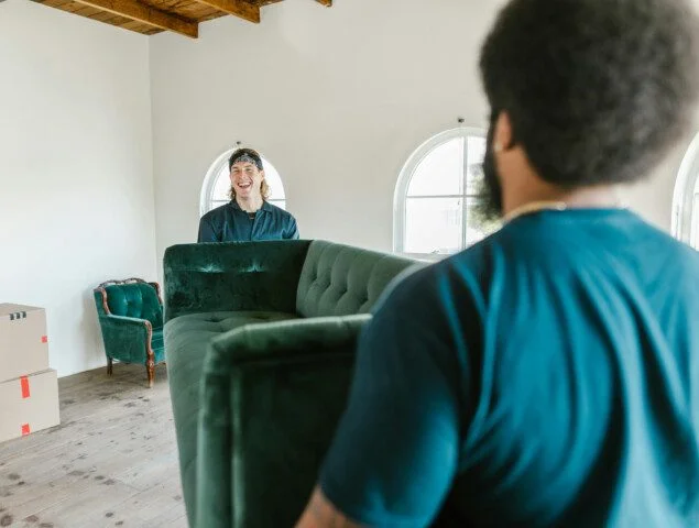 Two people carrying a green sofa in a bright room with boxes.
