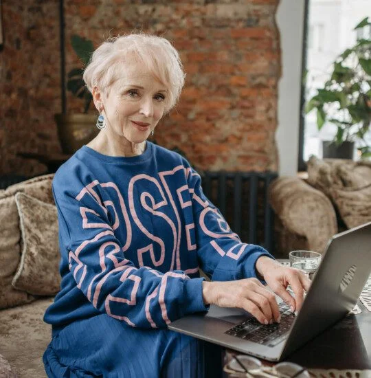 Older woman in blue sweater typing on a laptop in a cozy living room.