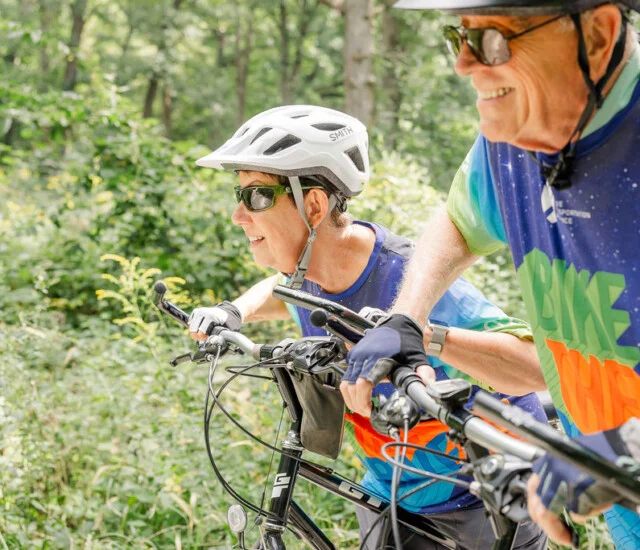 Two cyclists smile on forest trail with helmets and colorful jerseys.