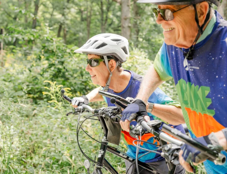 Two cyclists smile on forest trail with helmets and colorful jerseys.