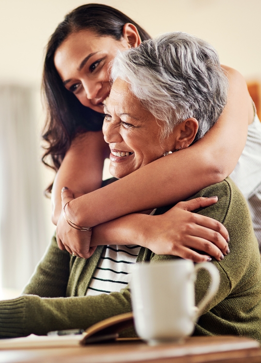 adult daughter hugs senior mother from behind while she's seated at a table