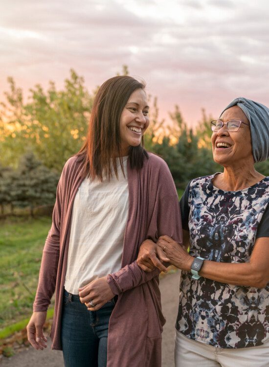 senior woman and her adult daughter walk arm-in-arm on a scenic walking path at sunset, smiling