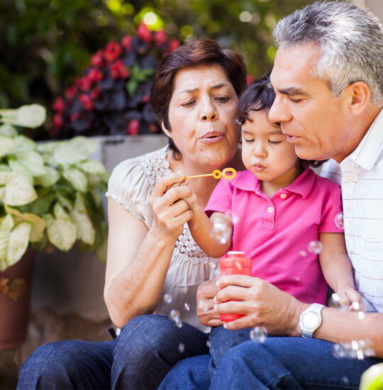 grandparents hold granddaughter in their laps and help her blow bubbles