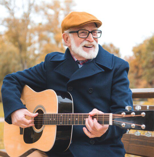 senior man seated on park bench in warm fall clothes smiles and plays acoustic guitar