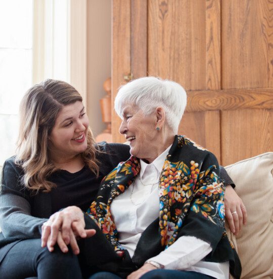 senior woman and adult daughter sit on a couch close together, smiling and holding hands