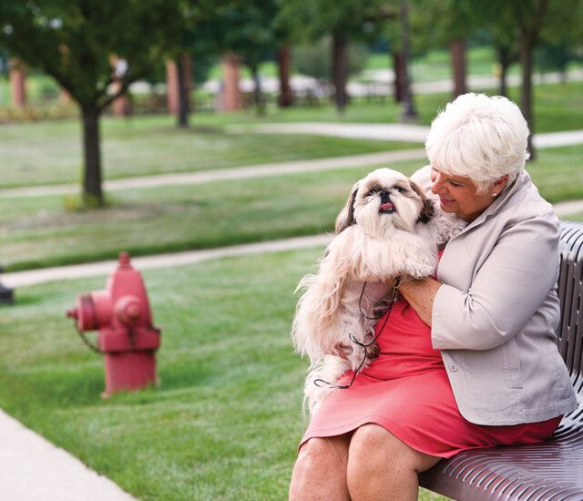 senior woman in dress sits on bench with her fluffy dog outside at Oak Trace Senior Living Community