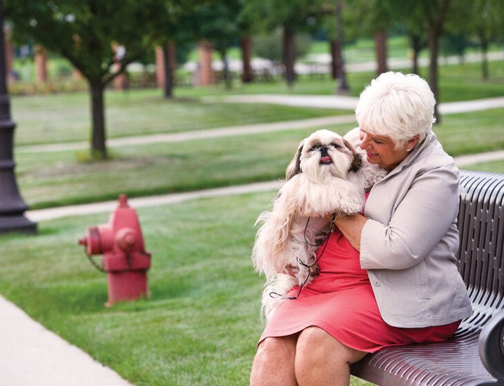 senior woman in dress sits on bench with her fluffy dog outside at Oak Trace Senior Living Community