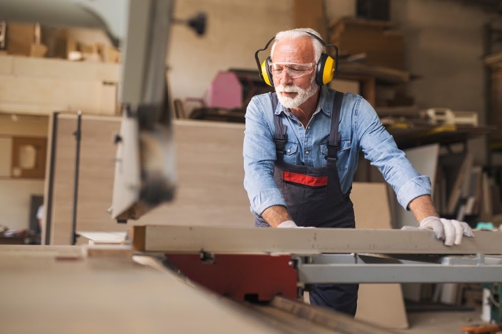 senior man wearing protective glasses and earmuffs while sawing piece of wood in his workshop