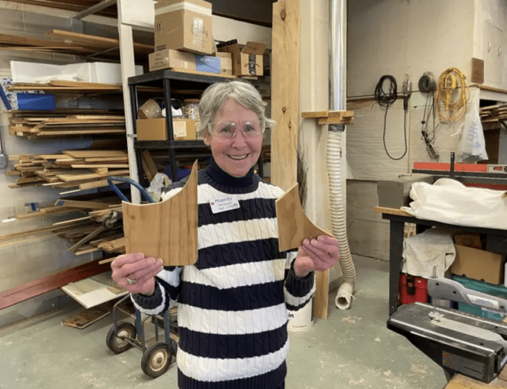 Oak Trace Senior Living resident Mandy Stewart smiles inside a workshop, wearing protective goggles and holding up pieces of wood