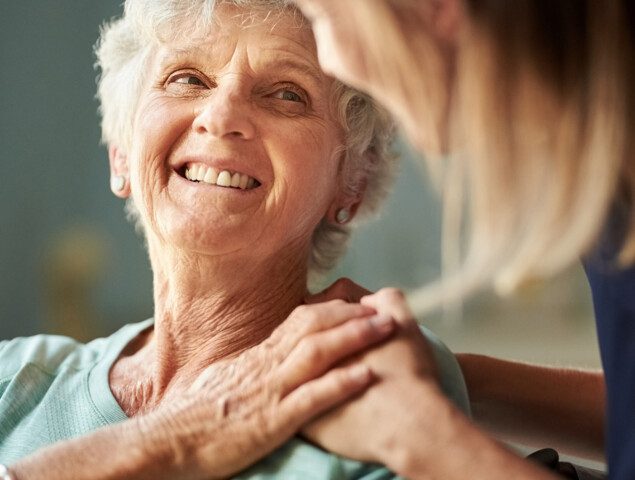 senior woman smiles up at caregiver, who gently places her hands on her shoulder