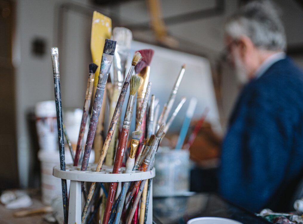 close-up of paintbrushes with senior man painting in the background