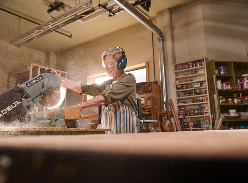 senior woman with protective glasses and headphones uses saw to complete woodworking project