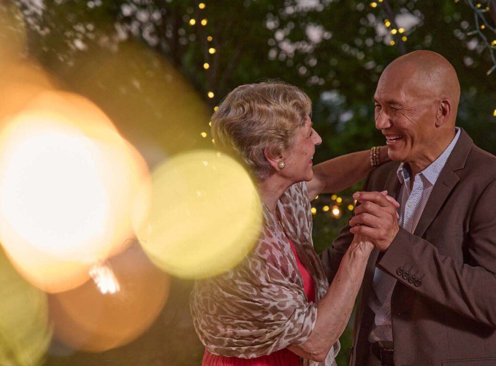 senior couple in elegant clothing dances together outside at sunset under fairy lights at Oak Trace Senior Living Community