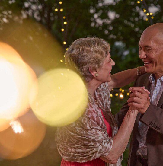 senior couple in elegant clothing dances together outside at sunset under fairy lights at Oak Trace Senior Living Community