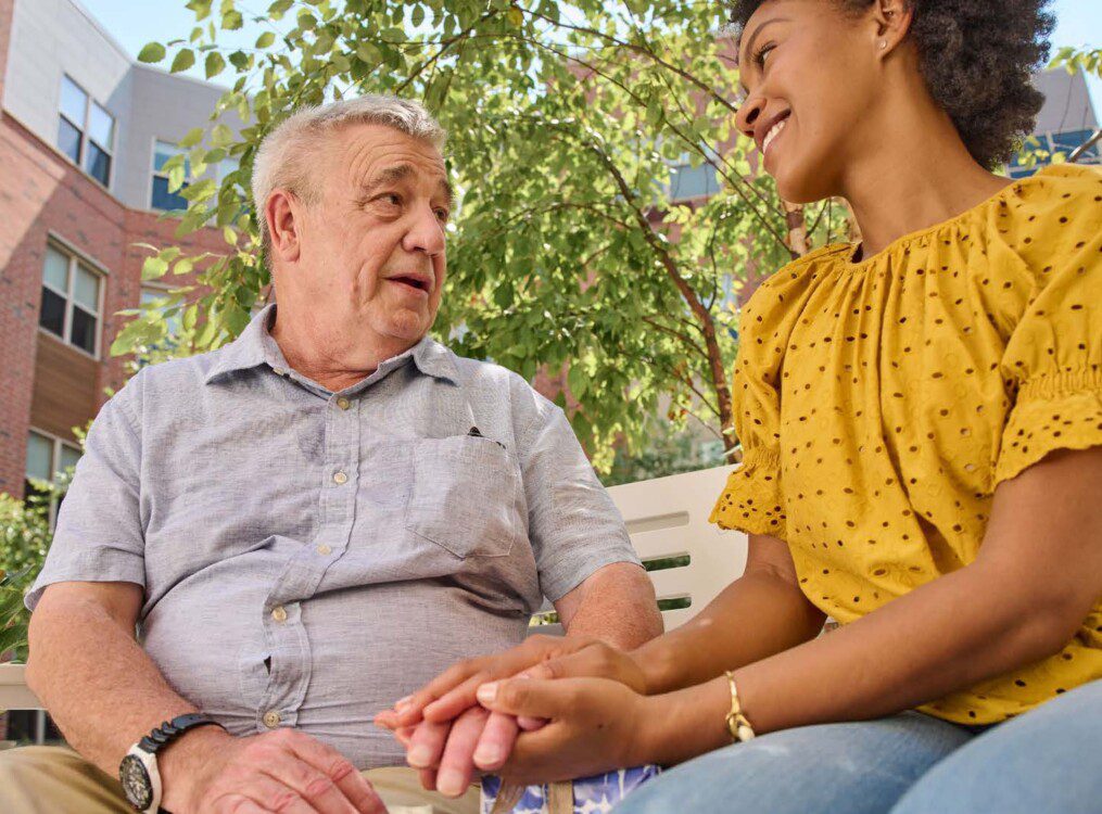 senior man chats with his adult daughter on a bench outside of Oak Trace Senior Living Community