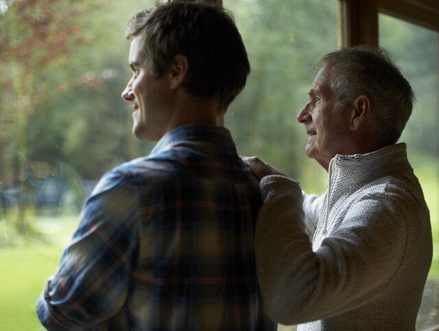 Senior father and adult son standing next to each other and looking out a window.