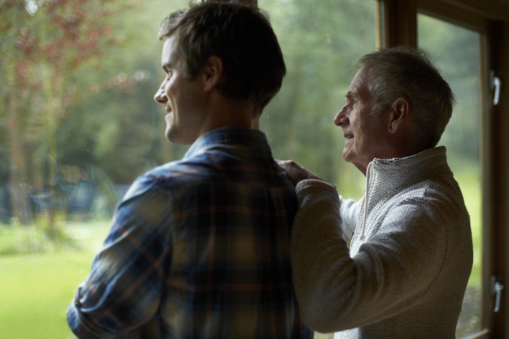 Senior father and adult son standing next to each other and looking out a window.