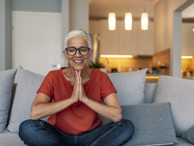 senior woman sits with her legs up, practicing meditation in her apartment at Oak Trace Senior Living Community