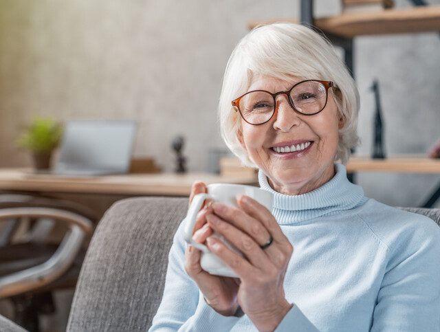senior woman seated on cozy chair smiles and holds coffee mug
