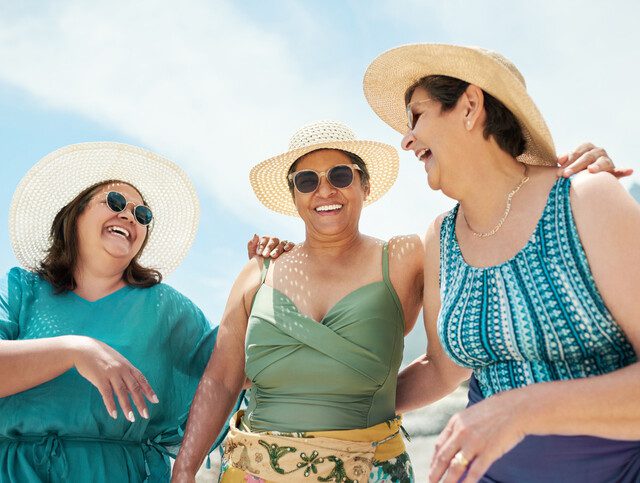 group of three senior women in bathing suits and stylish hats laugh together