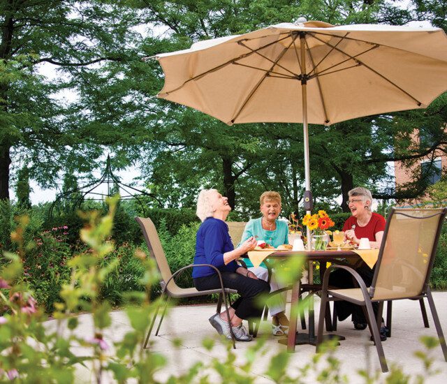 Group of three senior women laugh and enjoy brunch together outside in the garden area of Oak Trace Senior Living Community