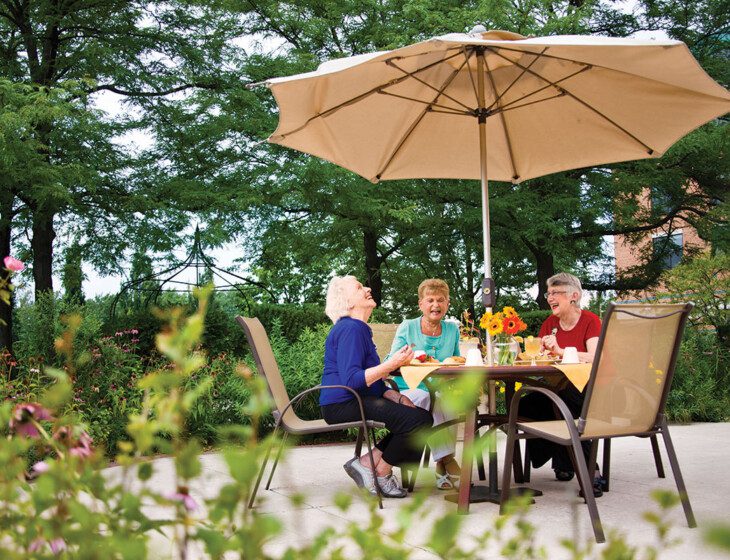 Group of three senior women laugh and enjoy brunch together outside in the garden area of Oak Trace Senior Living Community