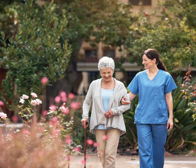 senior woman with cane smiles and walks through the garden with her caretaker at Oak Trace Senior Living Community