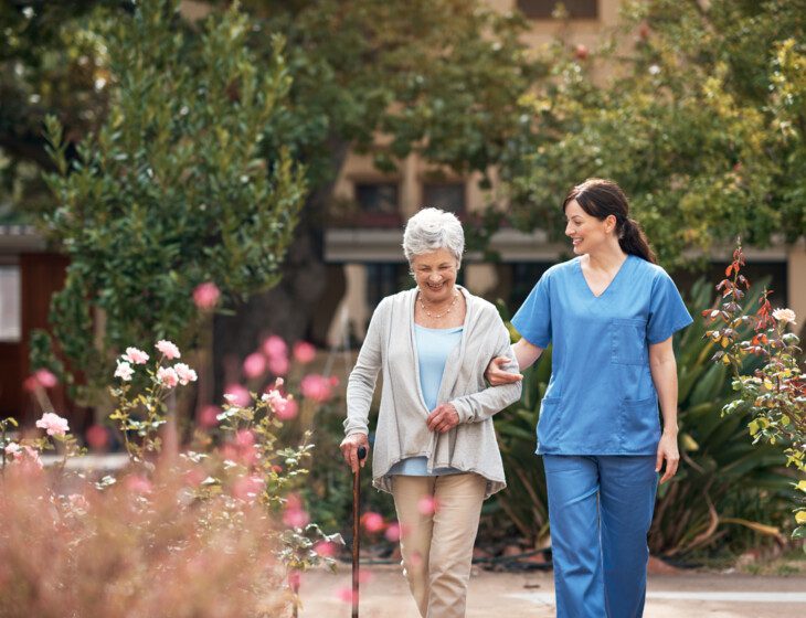 senior woman with cane smiles and walks through the garden with her caretaker at Oak Trace Senior Living Community