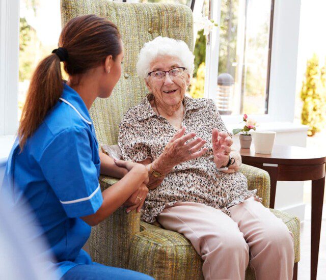 Senior woman seated in chair smiles and talks to her caretaker, who kneels beside her