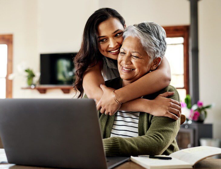 senior woman sits at laptop smiling while hugged from behind by her adult daughter