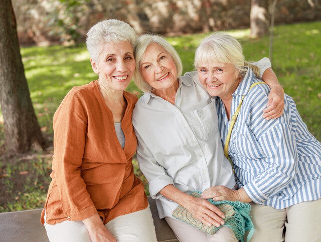 three senior women sit together outdoors, smiling and embracing