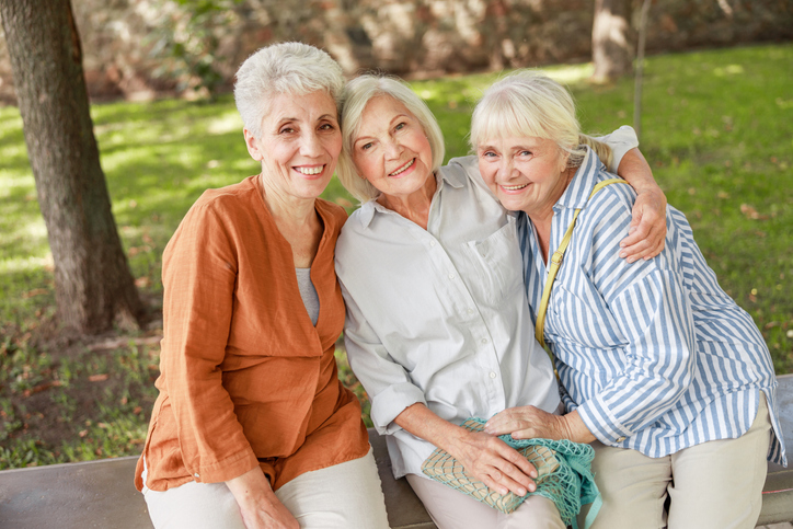 three senior women sit together outdoors, smiling and embracing