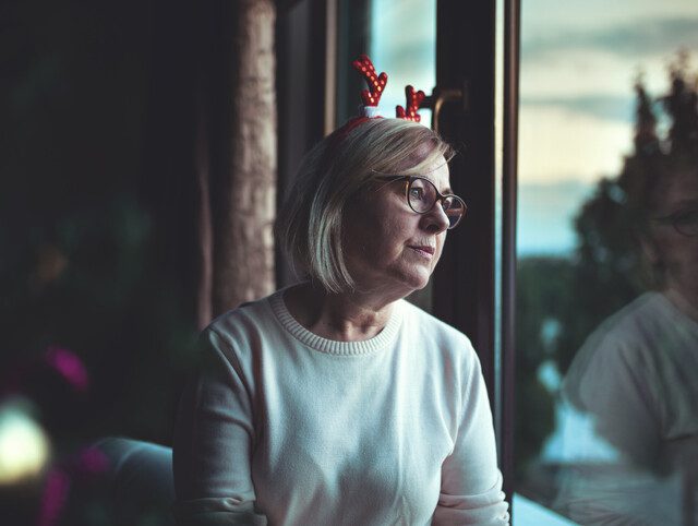 Senior woman wearing Christmas reindeer headband looks out of a window, lonely