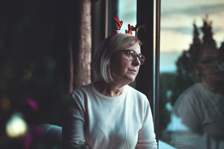 Senior woman wearing Christmas reindeer headband looks out of a window, lonely