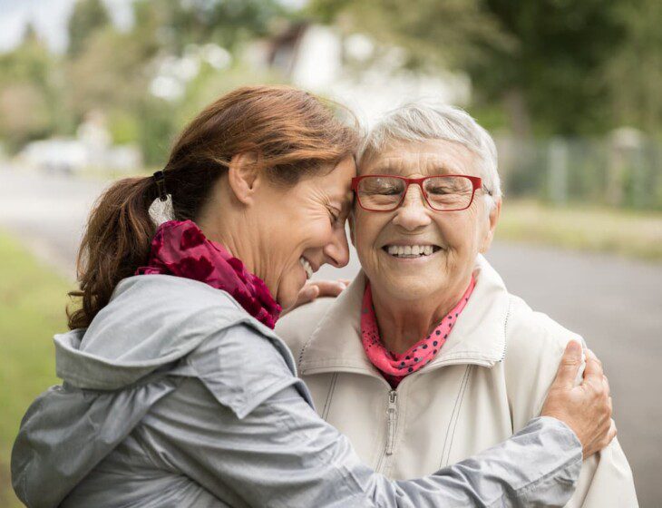 Senior woman and her adult daughter embrace for a hug on a walking path outside of Oak Trace Senior Living Community