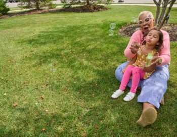 Shot 03 MinnardSkiles MiaPerez 0184 grandmother smiles and helps granddaughter blow bubbles while sitting on the grass outside
