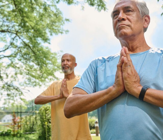 two senior men practice yoga poses and look off into the distance, backdropped by beautiful landscaping