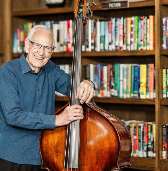 Elderly man smiling, holding a double bass, standing in a library.