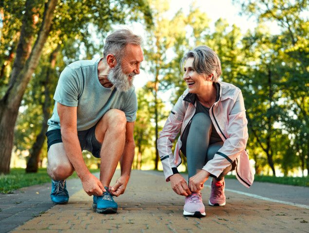 Older couple tying sneakers on a park path, smiling at each other.