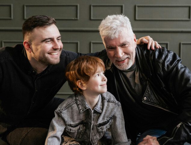 Three generations: grandfather, father, and son smiling together while playing with wooden blocks.